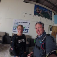 Mark and woman smile in front of Eugene Skydivers sign in the hangar